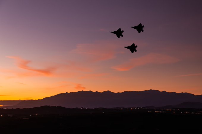 The image shows three airplanes flying high in the sky, creating an illusion of movement as they appear to be racing or competing with each other. The sun is setting behind these planes, casting a warm glow over their silhouettes against the horizon. This scene suggests that the airplanes might be participating in an air show or demonstration event at dusk, which often features aerobatic performances and impressive stunts for the audience's enjoyment.