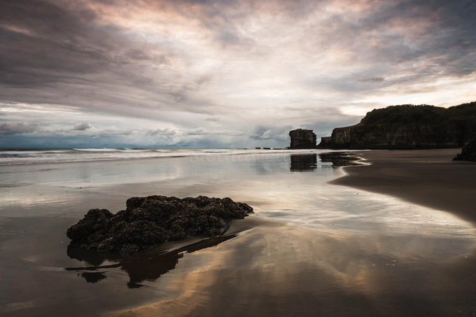 A dramatic coastal scene featuring a moody sky with sweeping cloud formations reflecting on the wet sandy beach, with rocky cliffs in the distance. This atmospheric image perfectly captures the essence of cloud photography, showcasing the interplay between the dynamic sky and the mirror-like shoreline at low tide.