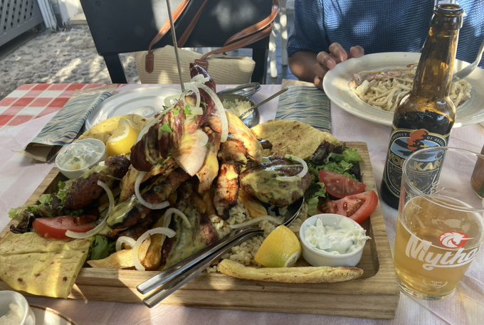 A wooden platter piled high with grilled chicken, lamb skewers, pita, rice, fresh veggies, and lemon wedges sits on a red-checkered table alongside a Mythos beer glass and a bottle—perfectly matching the "last supper" vibe of a hearty Greek family meal.