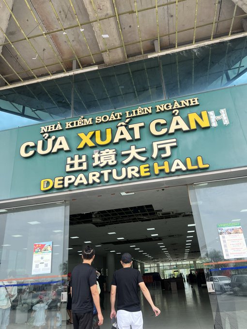 Two men in black shirts walk toward the entrance of a building marked 'CỬA XUẤT CẢNH' and 'DEPARTURE HALL'.
