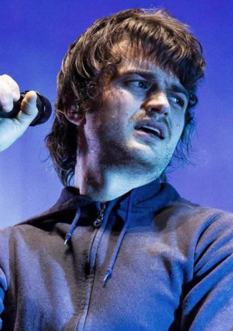 A performer with shaggy brown hair and a slight mustache sings into a microphone against a blue-lit background, likely Djo, the artist behind "End of the Beginning," which Polymarket traders predict has a 55% chance of becoming Spotify's top song of 2026.