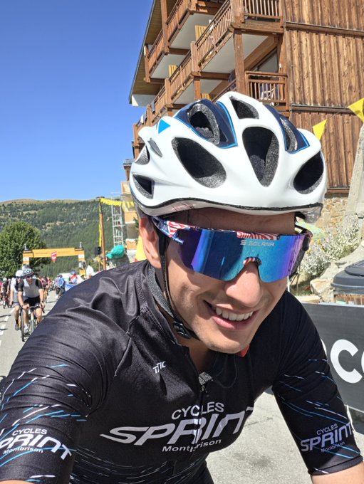 A close-up selfie of a smiling cyclist wearing a white helmet, blue mirrored sunglasses, and a black "CYCLES SPIN" jersey, riding in an outdoor event with wooden buildings behind him.