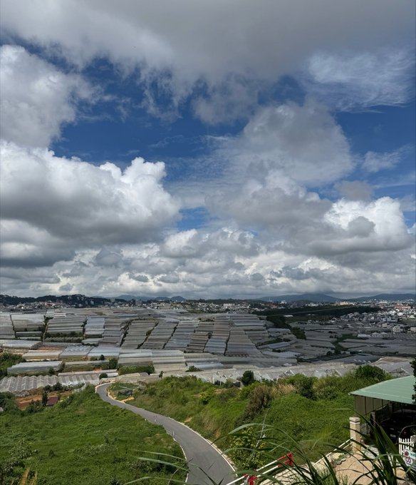 A scenic landscape photograph showing a vast expanse of greenhouses under a dramatic cloudy blue sky, with a winding road and distant mountains.