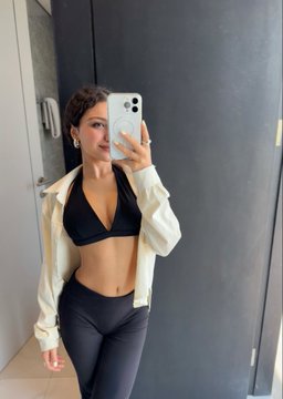 A woman takes a mirror selfie wearing a black bralette and leggings with a white jacket, showing her fit physique in what appears to be a gym or dressing room setting.