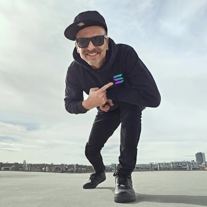 A man wearing a black cap, sunglasses, and a black hoodie with a colorful logo points at it while crouching on a rooftop against a cloudy sky.