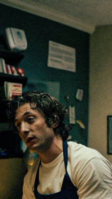A cinematic photo of a young man with curly hair wearing a white t-shirt and dark apron, looking to the side in what appears to be a kitchen setting.