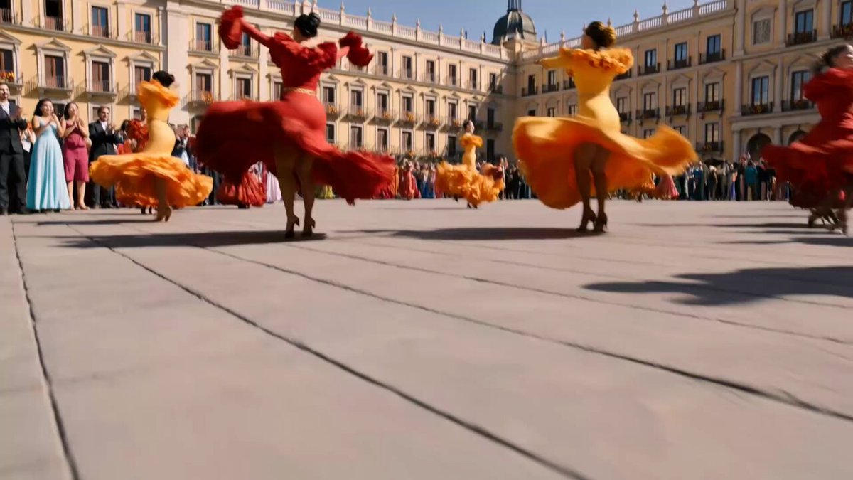 The image shows a group of people dancing in front of a building, possibly a palace or a church. The dancers are wearing colorful dresses that stand out against the backdrop. There are at least five visible figures in the scene, with one person on the left side and four others spread across the right side. They appear to be enjoying themselves as they dance together.