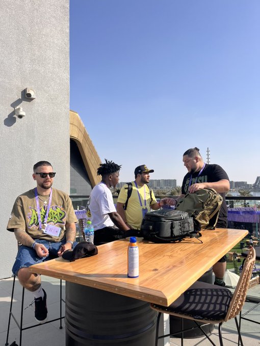 A group of four men are gathered around a high wooden table outdoors on a sunny day. One man in a black t-shirt is opening a green tactical bag while the others, wearing sunglasses and casual clothing, watch or look on.