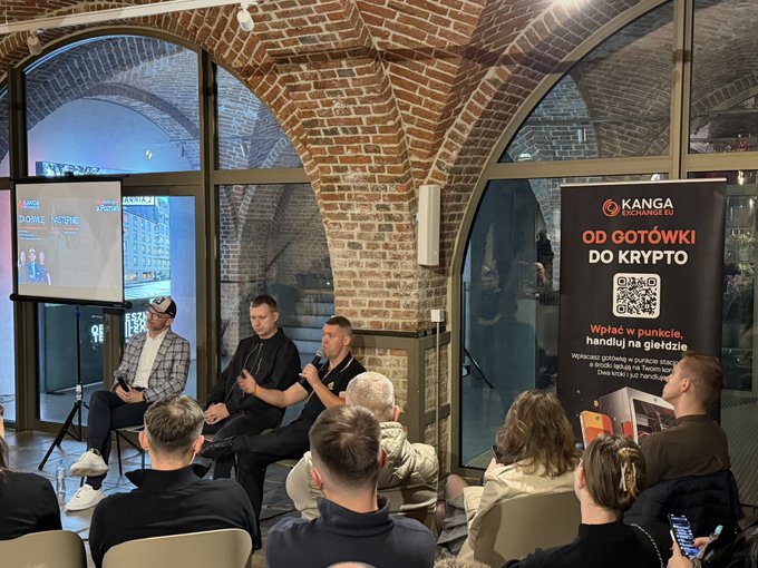 Three men sit on chairs in a brick-walled venue with arched windows, participating in a panel discussion with a banner reading 'KANGA EXCHANGE OD GOTÓWKI DO KRYPTO' beside them.