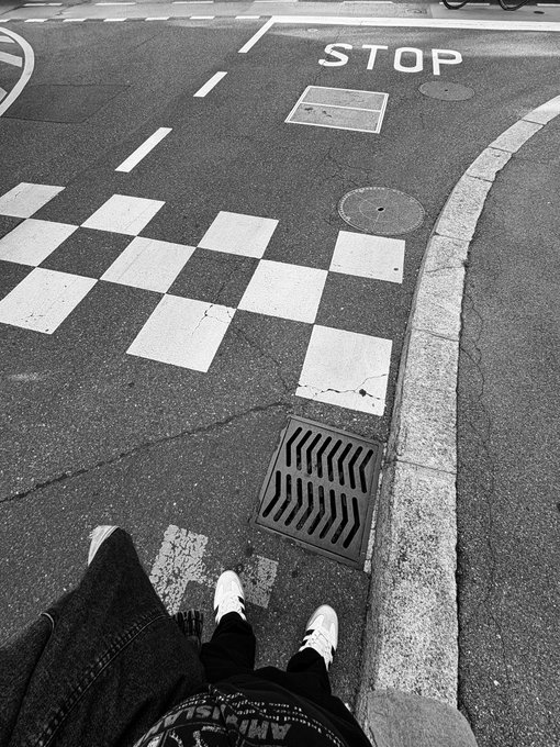 A black and white photo taken from above showing someone's feet in sneakers standing at a crosswalk with a checkered pattern, near a storm drain and 'STOP' road marking.