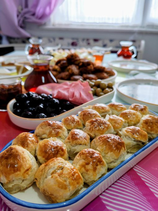 A traditional tea spread featuring a tray of golden-brown pastries (likely Turkish börek or similar), accompanied by black olives, green olives, dates, and glasses of Turkish tea.