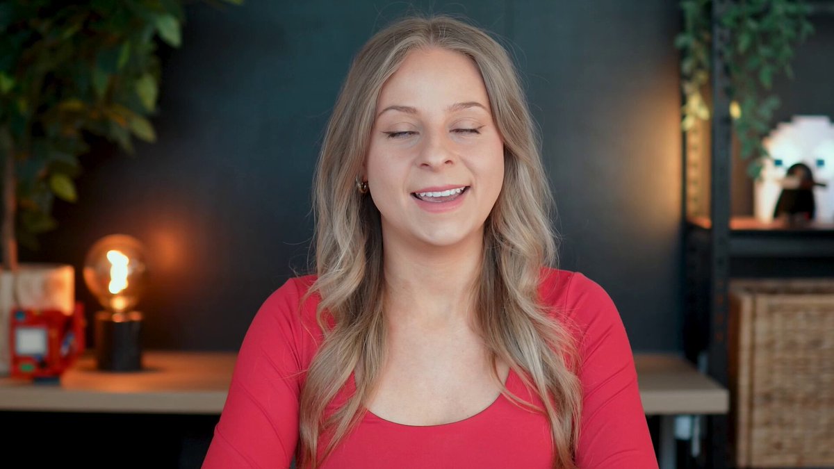 The image features a woman with blonde hair wearing a red top sitting at a desk or table. She appears to be smiling, possibly posing for a photo. The background of the image includes a green plant and a lamp, creating an indoor setting. There is also text in the image that reads "Cardano is becoming increasingly more connected to Bitcoin, Ethereum, Solana, Avalanche !" This suggests that the woman might be interested in or knowledgeable about cryptocurrency, as it seems to be related to her outfit or the environment she's in.