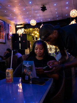 The image shows a man and woman sitting at a table, looking at a menu together. They are surrounded by various items such as a cell phone, a can of beer, and some other objects like a wallet and a book. The scene takes place in what appears to be a bar or restaurant setting with blue lights illuminating the area. The post seems to be promoting an event or gathering related to the German game "Caração 7-1" at a bar called "Nestle".