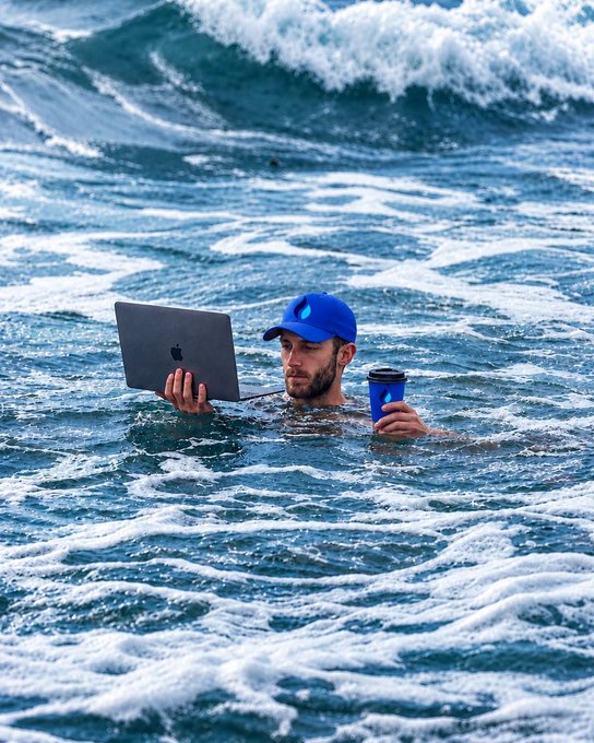 A man wearing a blue cap sits submerged in ocean waves, focused on his Apple laptop while holding a coffee cup, humorously taking "work from anywhere" to the extreme—likely trading stocks from somewhere far offshore.