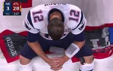 New England Patriots quarterback Tom Brady, wearing jersey #12, sits dejectedly on the bench with his head bowed during the third quarter of a game where Atlanta leads New England 28-3 with 7:47 remaining. The image of Brady in a moment of quiet defeat, captured under stadium lights against the backdrop of a "CHAMP" banner, perfectly embodies the "dark horse" sentiment—a champion brought low, humbled in a moment of unexpected vulnerability.