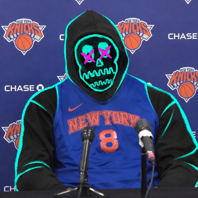 A person wearing a New York Knicks #8 jersey with a neon green and pink glowing skull mask and black hood, seated at a press conference with microphones in front of a Knicks-branded backdrop.