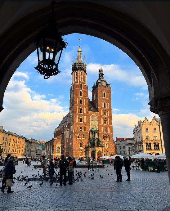 A view of St. Mary's Basilica in Krakow, Poland, framed through a stone archway with a hanging lantern, as people and pigeons gather in the square below.