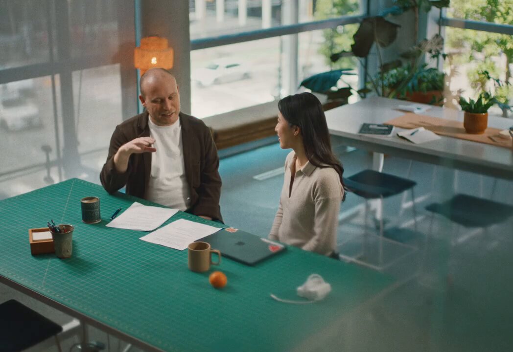 The image shows a man and woman sitting at a table, engaged in conversation. They are surrounded by various objects such as cups, books, and laptops. A laptop can be seen open on the table, suggesting that they might be discussing work or collaborating on a project. The scene takes place in an office setting with multiple chairs around the table, indicating a professional environment.