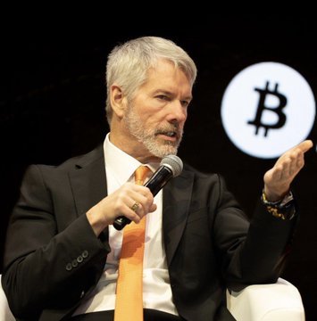 A gray-haired man in a dark suit and orange tie speaks into a microphone with a Bitcoin logo (₿) displayed behind him, appearing to be a featured speaker at a cryptocurrency event. The image aligns with the post's message about whale accumulation and long-term holding, as the speaker appears to be Michael Saylor, the prominent Bitcoin advocate known for his unwavering "never sell" stance referenced in the commentary.