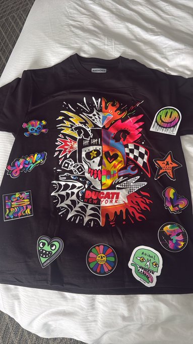 A black t-shirt decorated with various colorful patches and a large central graphic of a skull with flames and the text 'DUCATI NEW YORK', laid out on a white sheet.