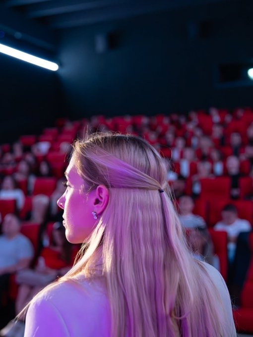 A young woman with long blonde hair stands in profile at a dimly lit conference venue with rows of red theater seats visible behind her. The image appears to be taken at a Bybit Europe crypto event, capturing an attendee amid the audience seating area.