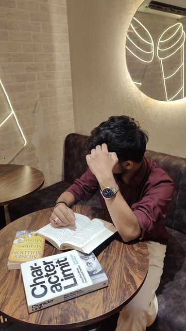 A young man in a maroon shirt sitting at a wooden cafe table with his hand on his forehead, reading books including 'Northis Singh' and 'Character Actor Limit' by Chetan Bhagat.