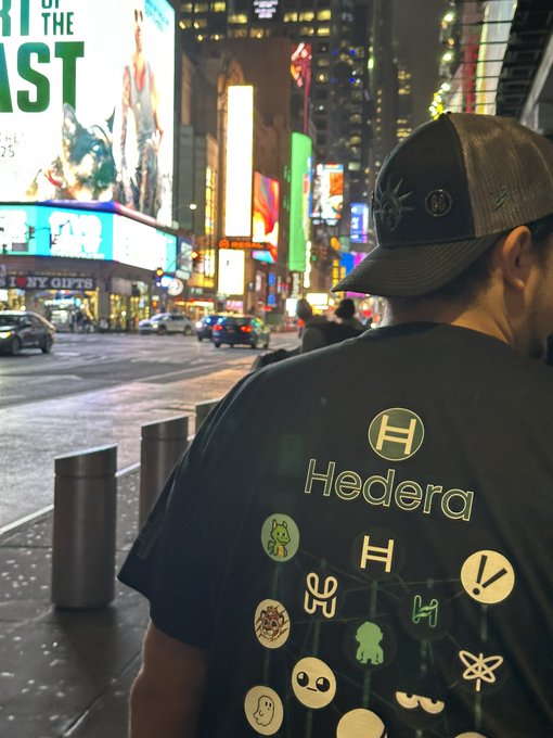 A person wearing a cap and a black 'Hedera' t-shirt with various logos stands on a Times Square street at night, facing the illuminated billboards.