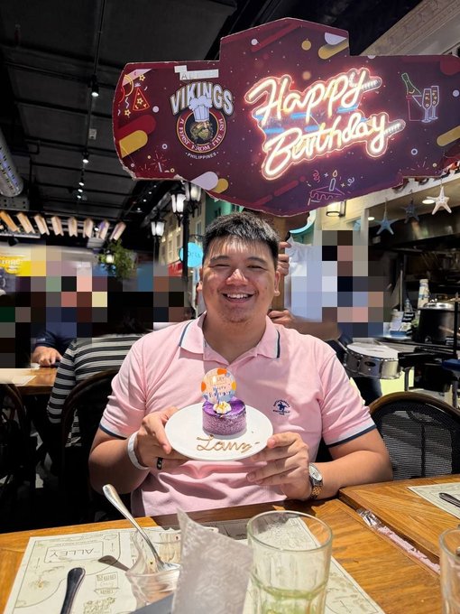 A man in a pink polo shirt smiling while holding a small birthday cake at a Vikings restaurant, with a 'Happy Birthday' banner and other patrons in the background.