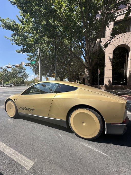 A gold-wrapped Tesla Cybertruck with "CYBERTRUCK" graffiti on its side is parked on a street corner.