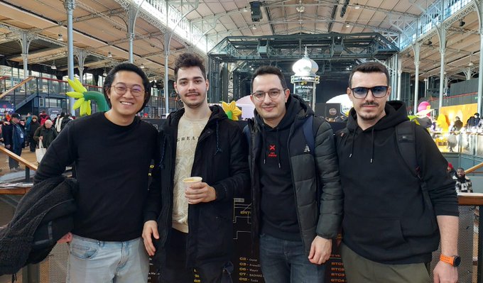 Four men pose together for a photo at what appears to be a trade show or convention, with a large hall featuring high ceilings and industrial decor in the background.