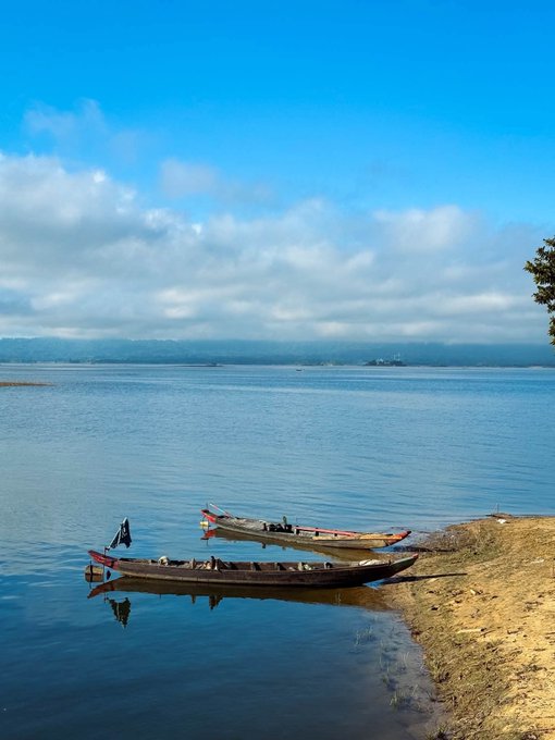 Two weathered wooden canoes rest gently at the water's edge of a calm lake, their reflections mirrored in the still blue surface beneath a soft, cloud-dappled sky. The serene scene evokes a moment of pause and reflection, echoing the contemplative tone of stepping into a new month—much like traders watching the market horizon for what September may bring.