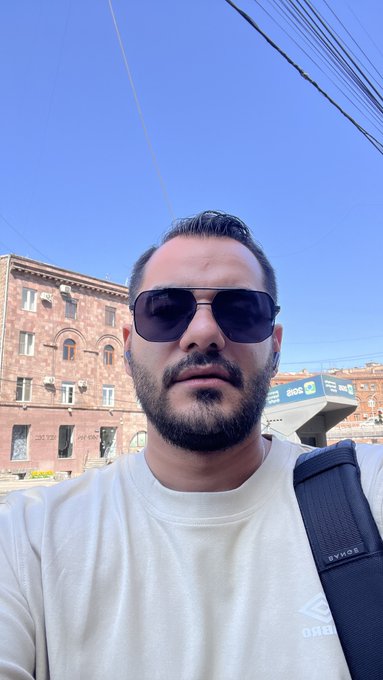A selfie of a bearded man wearing sunglasses and a white sweatshirt, standing on a sunny street with brick buildings in the background.