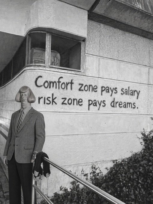 A black and white photo of a man in a checkered blazer and tie standing by a railing, with graffiti reading 'Comfort zone pays salary risk zone pays dreams.' on the wall behind him.