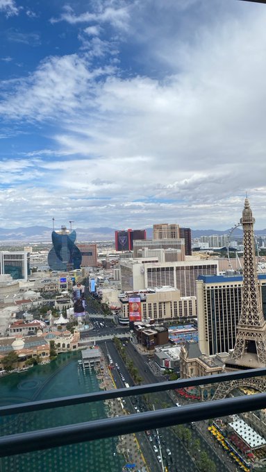 A daytime aerial view of the Las Vegas Strip from a high-rise balcony, featuring the Eiffel Tower replica, Caesars Palace, and other iconic hotels and casinos under a partly cloudy sky. "Gm" fits the sunny, optimistic morning vibe of waking up to this vibrant cityscape.