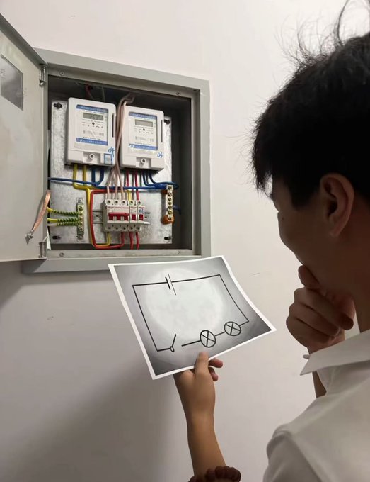 A young person in a white shirt stands before an electrical panel with meters, holding up a piece of paper showing a simple hand-drawn circuit diagram with a battery and light bulbs.