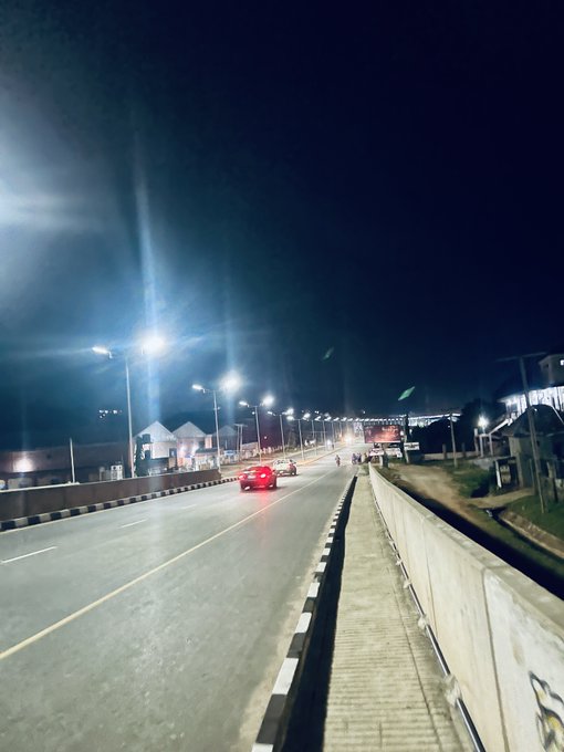 A nighttime photograph taken from a bridge or overpass looking down at a well-lit city street where a few cars are driving under bright streetlights.