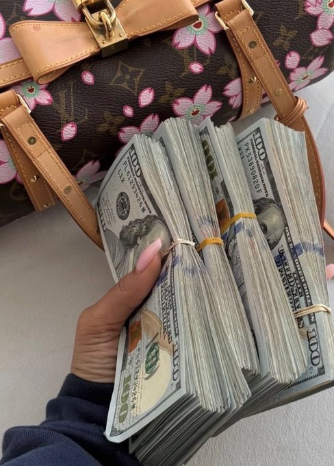 A hand holds several thick stacks of $100 US bills next to a Louis Vuitton monogram handbag decorated with pink floral patterns.