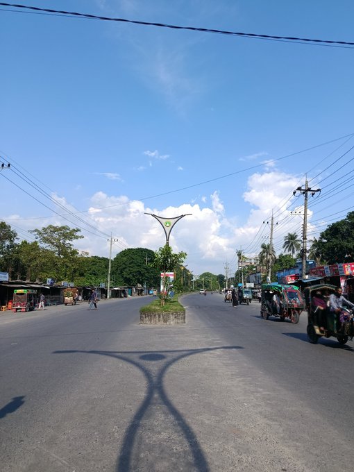 A street scene in Bangladesh with a central roundabout, a streetlight casting a Y-shaped shadow, and auto-rickshaws parked along the road under a blue sky.