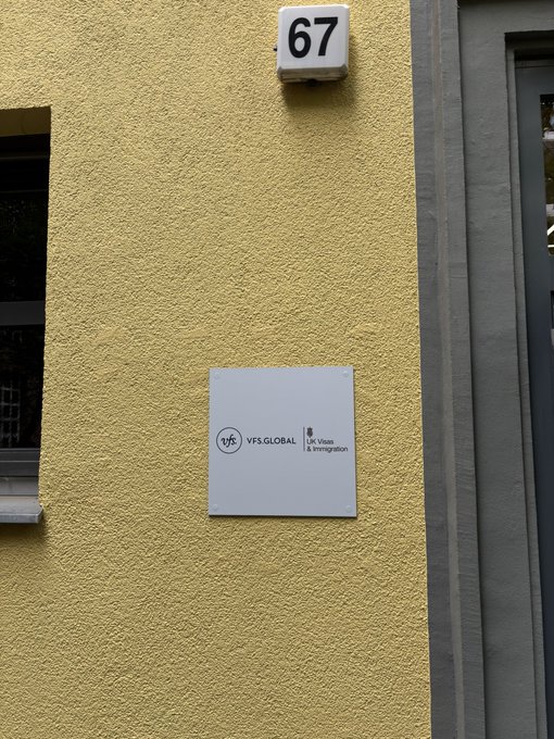 A photo of a textured yellow building exterior featuring a sign for 'VFS.GLOBAL' with text 'UK Visas & Immigration' below the number '67'.