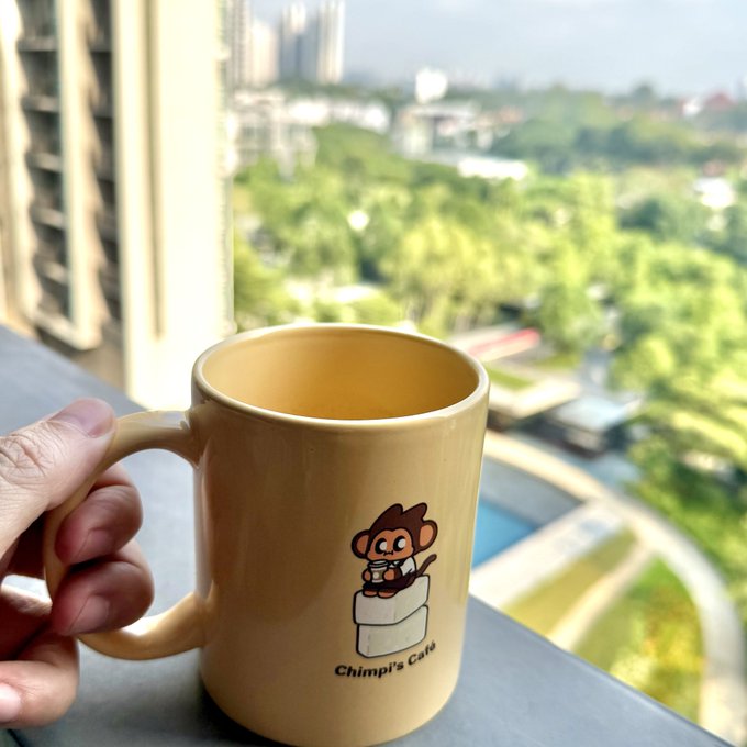 A hand holds a tan mug featuring a cartoon monkey and the text 'Chimpi's Cafe' on a balcony overlooking a lush green landscape.
