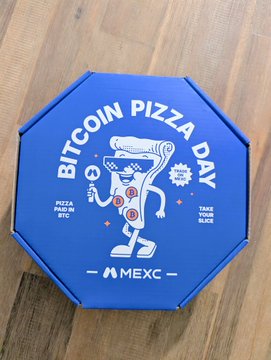The image features an open blue pizza box with a cartoon character of a pizza slice wearing sunglasses and holding a pizza cutlery set. The box has a message that reads "bitcoin pizza day" written on it, indicating a special occasion or promotion related to Bitcoin. There is also a small logo for the MXEC coin in the bottom right corner of the box.
