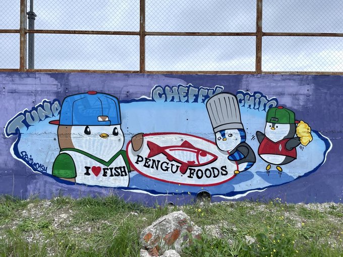 A colorful outdoor mural by artist @BUBOOST painted on a wall, featuring cartoon penguins wearing baseball caps and chef hats advertising "PENGU FOODS" and "I ❤ FISH."