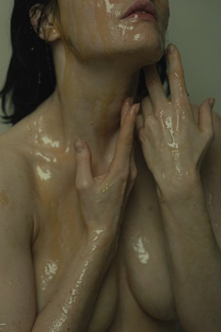 A vertical portrait captures a person covered in a viscous, amber-colored liquid, with their hands touching their neck and face. The image is a highly detailed artistic photograph that the accompanying post describes as "spectacular" work by @mindal_nft.