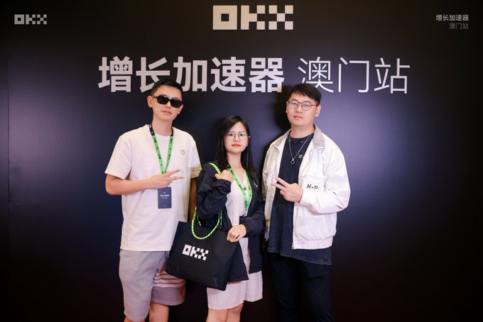 Three young people pose for a photo in front of a dark wall with 'OKX' and Chinese text '增长加速器 澳门站' (Growth Accelerator Macau Station).