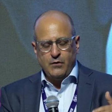 A man with glasses wearing a dark blazer and light blue shirt speaking into a microphone with his eyes closed.