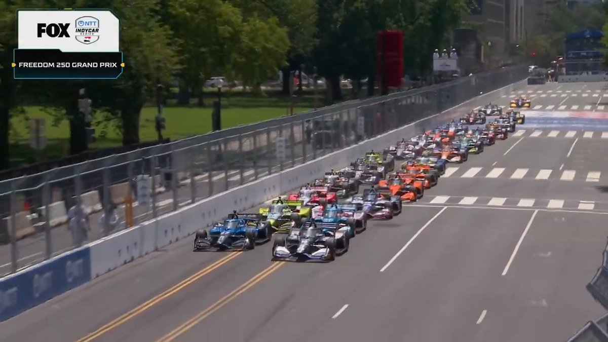 A FOX broadcast of the NTT INDYCAR Series Freedom 250 Grand Prix shows a pack of race cars speeding down a city street lined with trees.