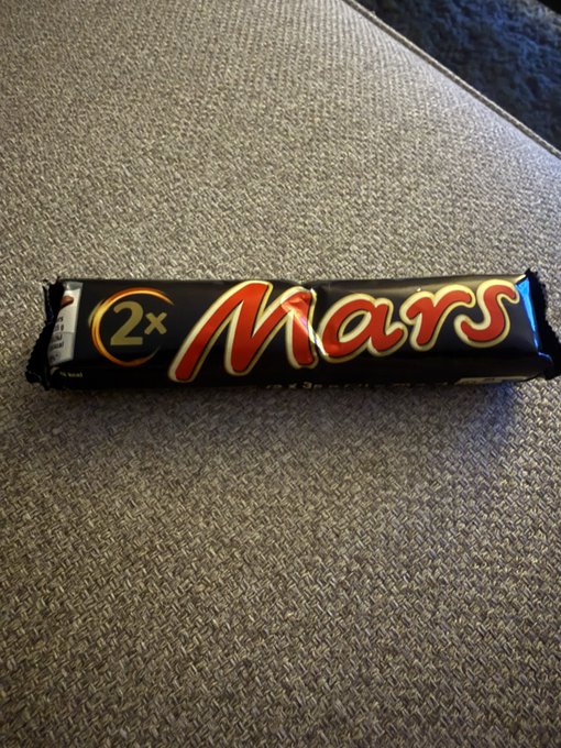 A 2x Mars chocolate bar wrapper (black packaging with red Mars logo) lying on a textured gray fabric surface.