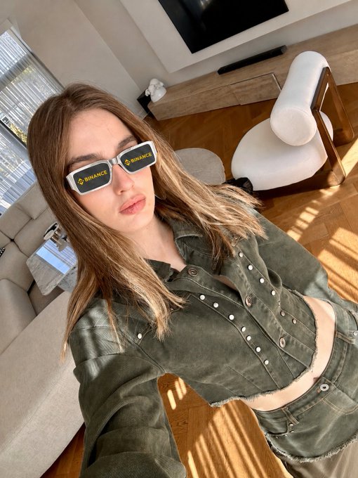 A woman takes a selfie wearing white sunglasses branded with the "BINANCE" logo in yellow, posing in a modern living room. The image ties directly to her post about Binance concerns, visually representing her connection to the exchange amid the news.