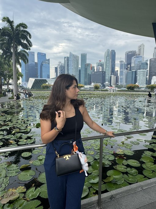 A woman in a black top and blue jeans stands by a pond with lily pads, holding a black handbag, with a modern city skyline in the background.