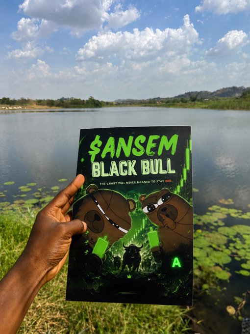 A hand holding a book titled '$ANSEM BLACK BULL - The chart has never meant to stay RED' featuring cartoon bears on the cover, photographed against a scenic lake background.